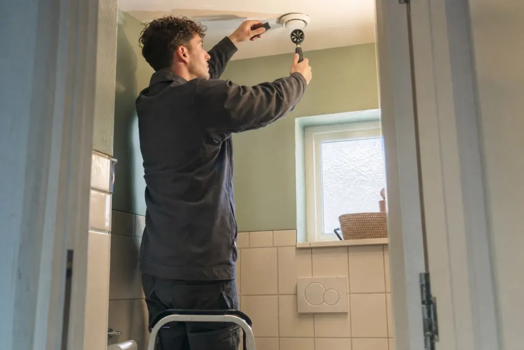 In het toilet staat een monteur op een opstapje en houdt een kleine windmeter en een zaklamp bij het afzuigventiel in het plafond
